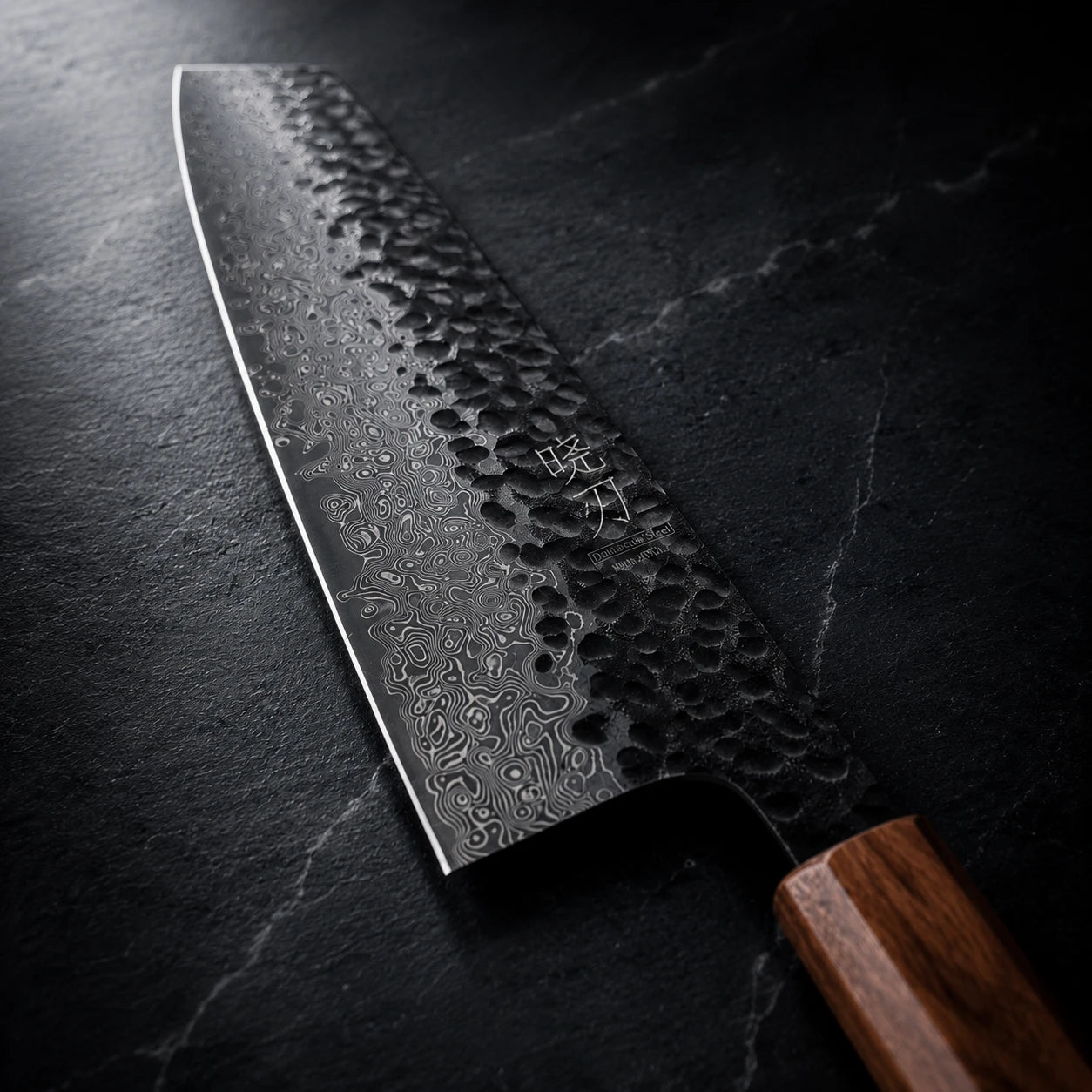 A Japanese-style kitchen knife with a hammered Damascus steel blade and a brown wooden handle, resting diagonally on a dark textured slate surface.