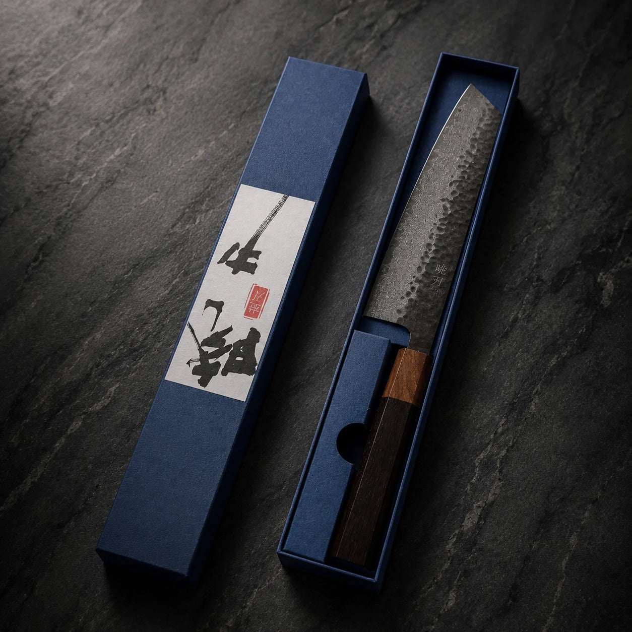 A Japanese-style chef's knife with a hammered steel blade and dark wooden handle, displayed in an open blue presentation box next to its matching blue box lid on a dark textured surface.