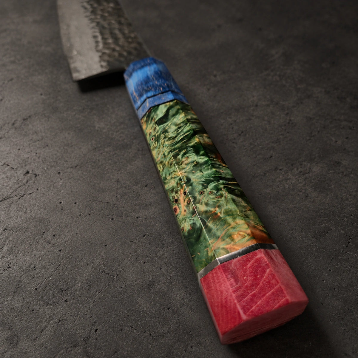 A close-up of a handmade knife with a textured dark steel blade, a blue wrapped section, and a marbled handle featuring green, orange, and red inlay, resting on a dark gray textured surface.