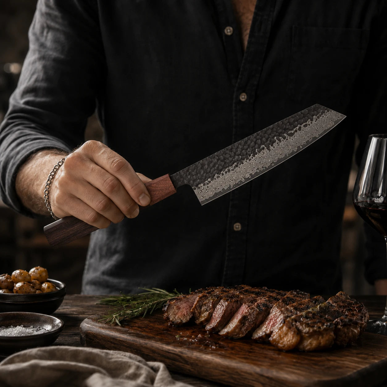 A person in a black button-up shirt holds a large chef's knife with a hammered Damascus-style blade and reddish-brown wooden handle over a wooden cutting board with sliced steak, rosemary, and small bowls of food.