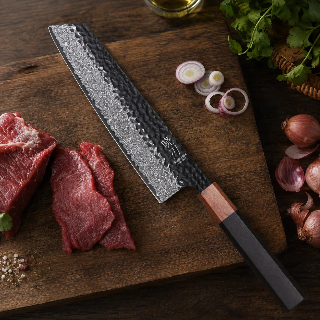 A Japanese-style kitchen knife with a hammered Damascus steel blade and a black handle with a copper-colored bolster, resting on a wooden cutting board surrounded by raw beef, red onions, garlic, and herbs.