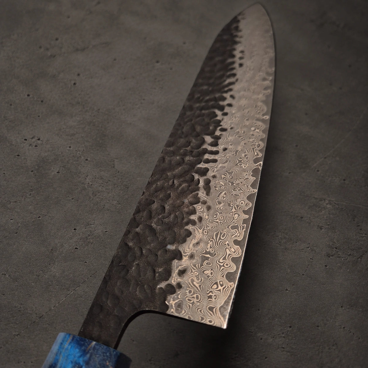 A Japanese-style kitchen knife with a hammered Damascus steel blade and a blue marbled handle, resting on a dark textured surface.