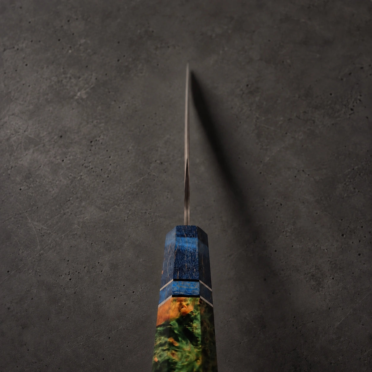 A close-up of a knife with a long, slender steel blade and a highly detailed, multicolored inlay handle. The handle features a textured blue top section and a lower section painted with a scenic landscape of green trees, orange foliage, and brown earth, set against a dark gray textured background.