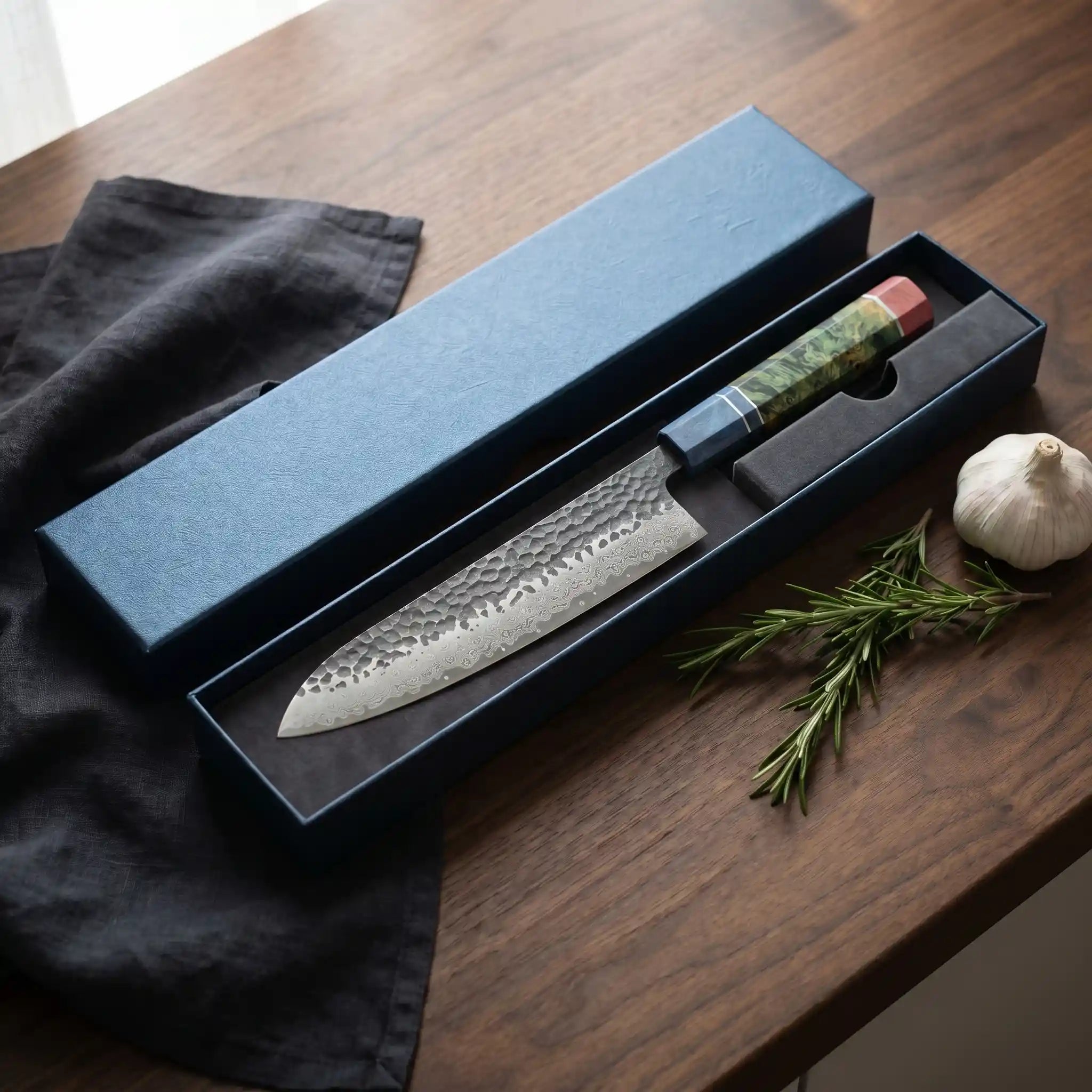 Japanese chef's knife with a textured handle in a black box on a wooden surface with herbs and garlic.