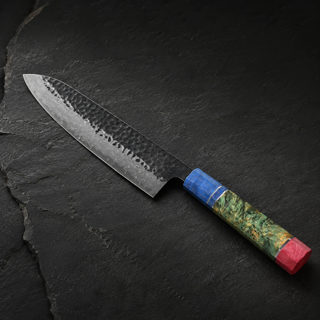 A Japanese-style kitchen knife with a hammered steel blade and a colorful handle featuring blue, green, yellow, and red sections, resting on a dark slate background.